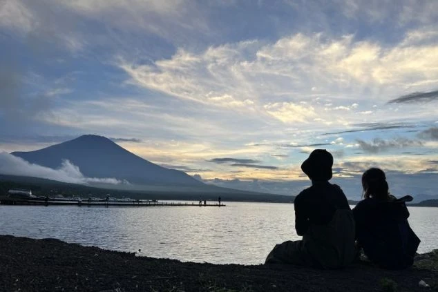 夕暮れ時の山中湖のほとりで、富士山を眺めながらリラックスするリゾートバイトスタッフの背中。休日に絶景を楽しめる、山梨県富士五湖エリアの住み込みバイト生活。