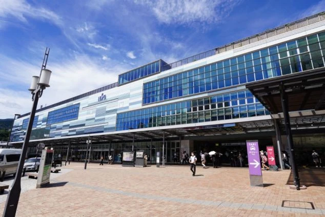晴天の下に建つ、モダンなガラス張りの大きな駅舎（熱海駅）。駅前広場は広々としており、タクシー乗り場の案内看板や数人の歩行者が写っています。