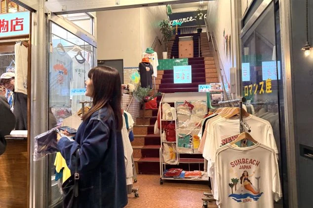 レトロな雰囲気の商店街にあるアパレルショップ。店頭には「SUNBEACH JAPAN ATAMI」とプリントされたTシャツなどが並び、奥には赤い絨毯が敷かれた階段が続いています。