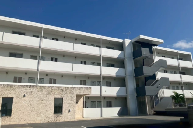 白い壁のアパート寮の外観。目の前には広々とした駐車場があり、自家用車やレンタカーを停められる。