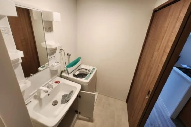 洗面台と洗濯機が置かれた、白い壁と木目調の扉があるシンプルな洗面所。