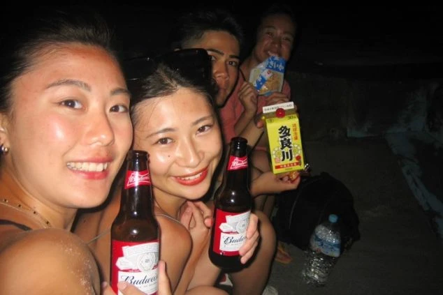 夜の屋外で、ビール瓶や紙パックのお酒、お菓子を手に持ちながら笑顔で自撮りをする男女4人のグループ。