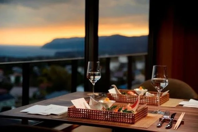 夕焼けに染まる海と山を望むレストランのテーブル席。竹細工の器に盛られた美しい前菜と、水が入ったワイングラスが並んでいる。