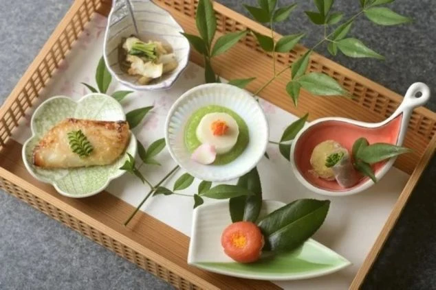 竹細工のトレイに、色とりどりの小皿が並んだ和食の盛り合わせ。焼き魚や季節の野菜などが、南天の葉を添えて上品に配置されている。58文字