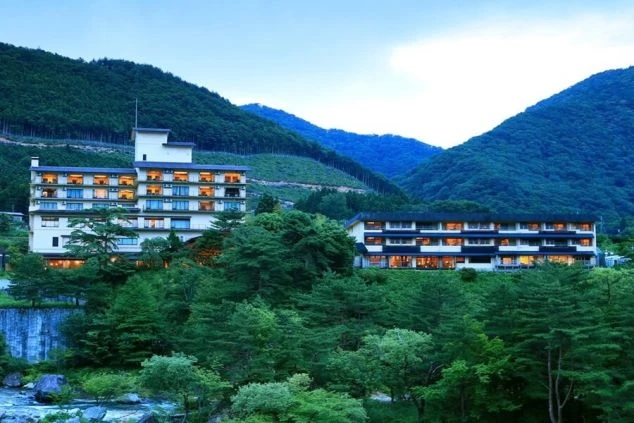 深い緑の山々と清流に包まれ、夜の明かりが美しく灯る自然豊かな温泉旅館の全景