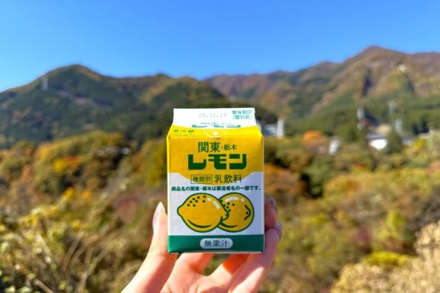 紅葉する山々を背景に、栃木のご当地飲料「レモン牛乳」を手に持つスタッフの日常