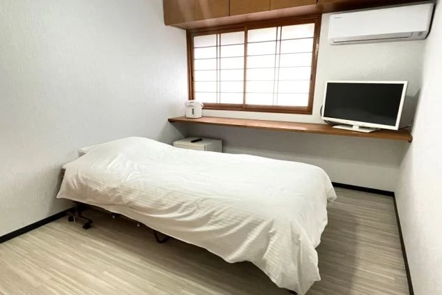 白を基調とした個室。白いシーツのシングルベッドがあり、窓際の木製カウンターにはテレビ、電気ポット、エアコンが設置されている。