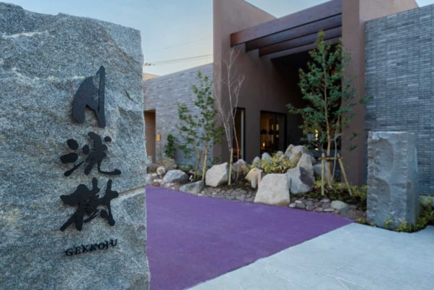 施設へと続くアプローチ。手前には店名が刻まれた大きな石碑があり、奥にはモダンな平屋建ての建物と植栽が並んでいる。