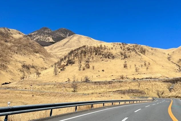 抜けるような青空の下、緩やかにカーブする道路の先に広がる、冬枯れした黄金色の山々と険しい岩肌の山頂。