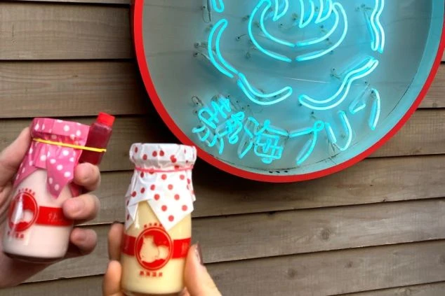 青いネオン看板を背景に、水玉模様の紙が被せられた2本の瓶入りプリンを手に持って並べている様子。