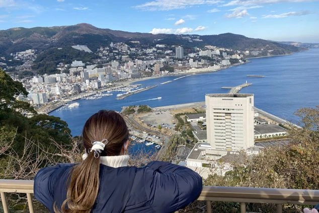 展望台の手すりに寄りかかり、眼下に広がる港町と青い海を眺める女性の後ろ姿。遠くには山々に囲まれた市街地が見える。