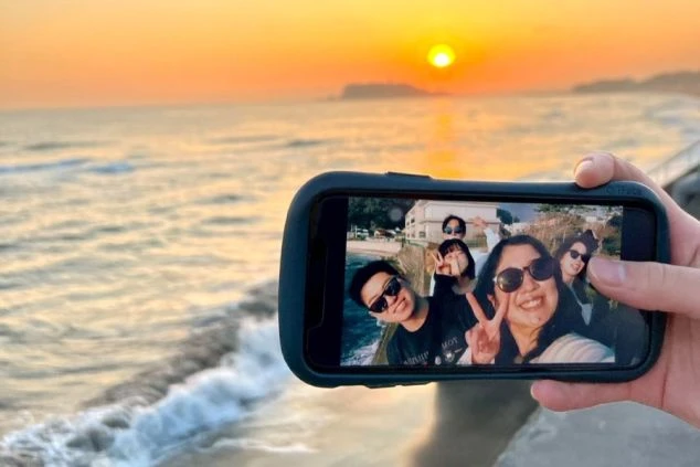 夕日が沈む海岸を背景に、スマートフォンの画面越しに友人たちとの自撮り写真を写している様子。海辺には穏やかな波が打ち寄せている。
