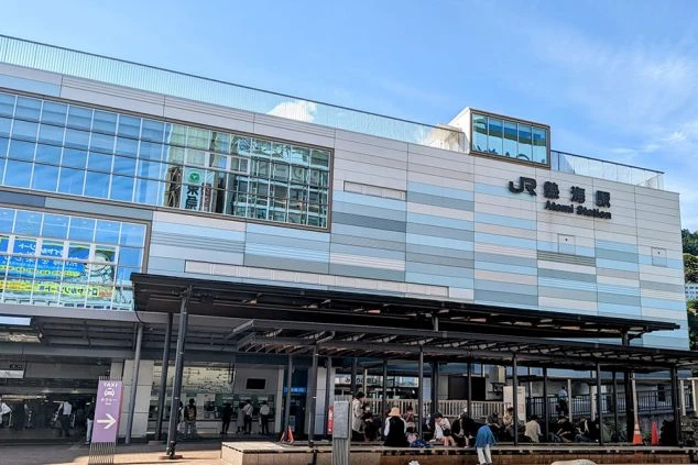 青空の下に立つ、現代的なデザインの大きな駅舎の外観。ガラス張りの壁面が特徴的で、手前の広場には屋根付きのベンチがあり、人々が休憩している。