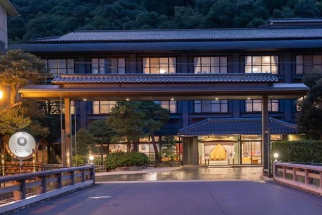 夕暮れ時の和風ホテルの外観。大きな門構えと、暖色の光が漏れる客室が並ぶ多層階の建物。手前には橋があり、落ち着いた情緒ある佇まい。