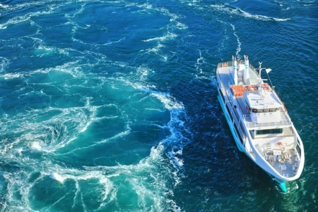 激しく渦を巻く青い海面を、白い観光遊覧船がゆっくりと進んでいる様子を高い位置から捉えた写真。