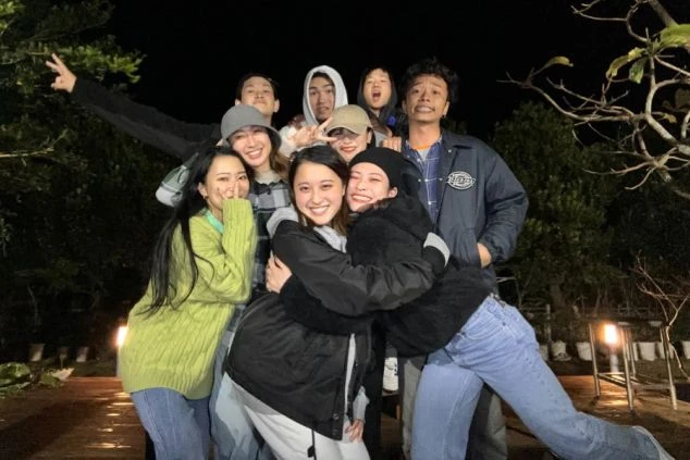 夜の屋外で、男女8人のグループが笑顔で肩を組んだり抱き合ったりしながら、仲睦まじく集合写真を撮っている様子。