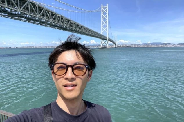 青空の下、海の上に架かる巨大な白い吊り橋を背景に、サングラスをかけた男性が自撮りをした写真。