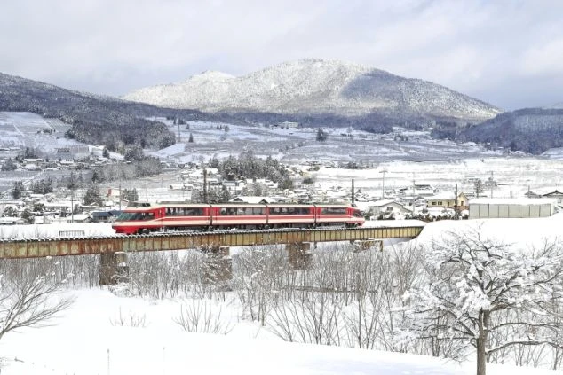一面の銀世界の中、鉄橋を渡る赤と白の特急列車。背景には雪化粧をした山々と、静かに佇む冬の集落が広がっている。