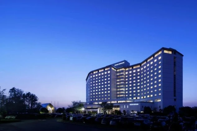 夕暮れの紺色の空に映える、明かりの灯った近代的な高層ホテルの外観。手前の駐車場には多くの車が停まっている。