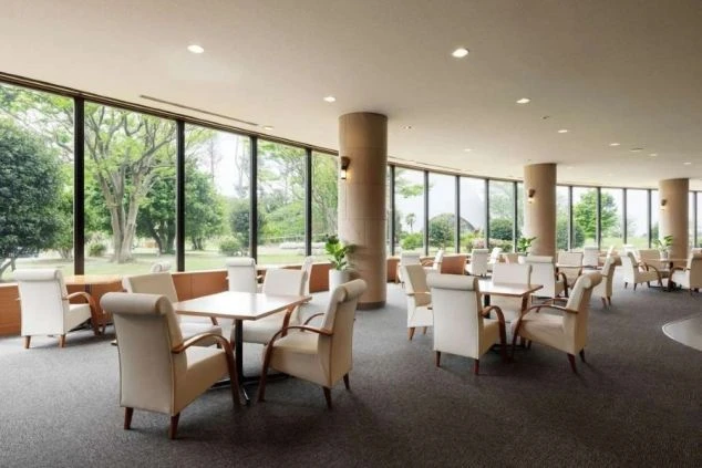 大きな曲線状の窓から明るい光が差し込む、広々としたラウンジ。白い椅子と木製のテーブルが並び、外には豊かな緑が見える。