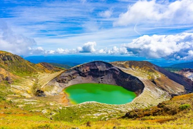 荒々しい火口壁に囲まれた、鮮やかなエメラルドグリーンの円形火口湖。青い空と白い雲が広がる、神秘的な高山の風景。