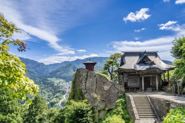 切り立った岩場の上に建つ古いお堂。晴天の下、眼下には緑豊かな山々と集落が広がり、遠くの連峰まで見渡せる絶景。