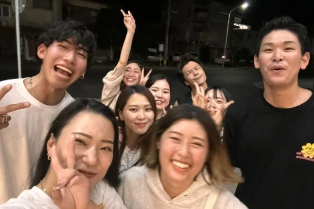 夜の屋外で、男女10人ほどのグループが笑顔ではしゃぎながら撮影した自撮り写真。皆でピースサインなどを送り合っている。