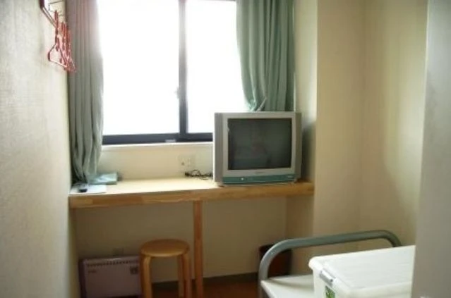 窓際の木製デスクに置かれた据え置き型のテレビ。窓には薄緑色のカーテンがかかり、デスクの下には木製の丸椅子が置かれている。