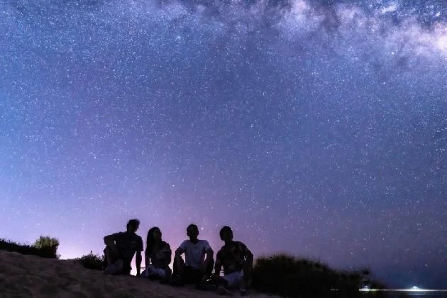 満天の星空の下、砂浜に座って空を見上げる4人の人物のシルエットの画像。天の川が輝き、都会を離れて体験できる、忘れられない夜の絶景と仲間との特別な時間を表現している。