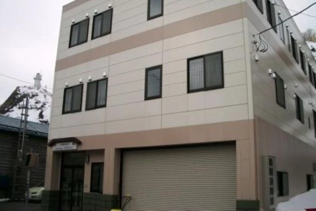 白とベージュのパネル張りの3階建てアパートの外観。1階には大きなシャッター付きのガレージがあり、背景の山には雪が積もっている。