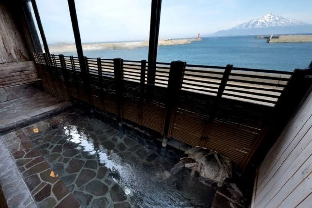 石造りの内湯。大きな窓からは穏やかな海が広がり、遠くには雪を頂いた美しい独立峰がそびえ立っている。木製の格子が窓辺に配され、情緒ある風景。