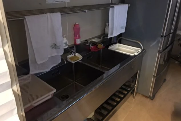 共用のステンレス製流し台。2つのシンクがあり、上部にはタオル掛けが設置されている。横には大型の冷蔵庫が置かれている。