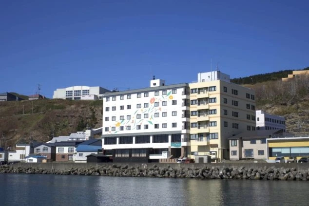 穏やかな海沿いに建つ多層階のホテル。白い外壁にはオレンジや黄色の花々のイラストが描かれ、隣にはベージュ色の建物が並んでいる。背後には緑豊かな丘が広がっている。