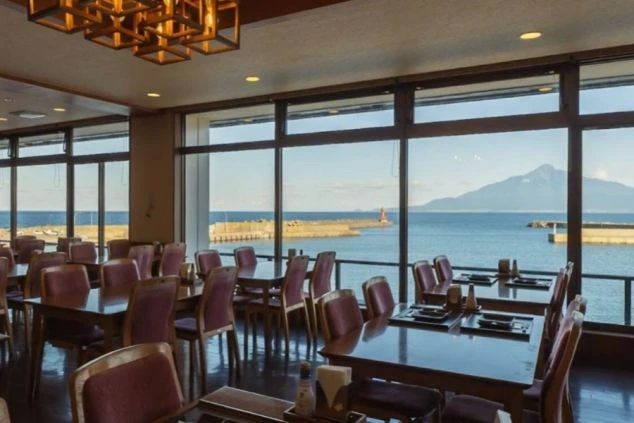 大きな窓から青い海と山を一望できる、開放感あふれるレストラン。木製のテーブルと紫色の椅子が並び、天井にはモダンな格子状の照明が設置されている。