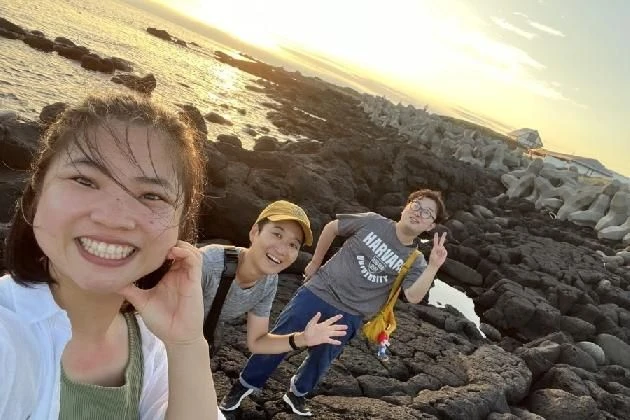 ワクトリでリゾートバイトしたスタッフが撮影した、北海道礼文島の夕焼けに染まる海岸で、笑顔でポーズをとる3人の若い男女の画像。岩場とテトラポッドが見える背景は、礼文島ならではの自然景観。