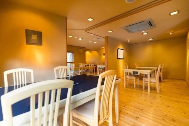 明るい木目調の床と温かみのある壁に囲まれた、清潔感のあるお食事処。等間隔に配置されたテーブルと椅子が並び、落ち着いて食事ができる和モダンな空間。