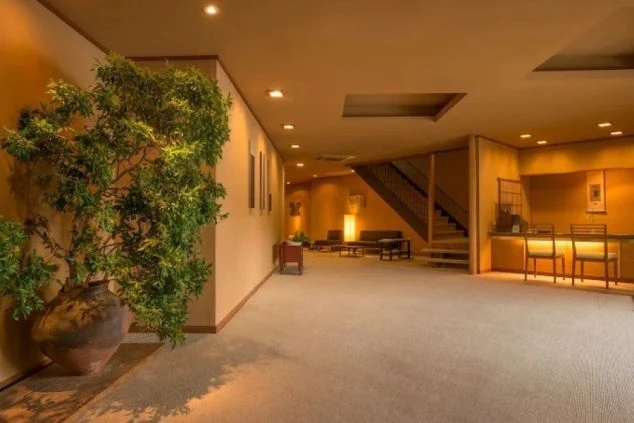 落ち着いた照明の広々とした旅館ロビー。手前には立派な盆栽が置かれ、奥にはフロントカウンターや2階へ続く階段、くつろげるソファスペースが整然と並んでいる。