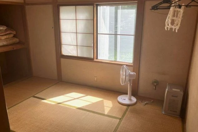 日当たりの良い畳敷きの和室。窓際には白い扇風機と小型のヒーターが置かれ、壁にはハンガーラック、奥には布団が収納された押し入れがある。