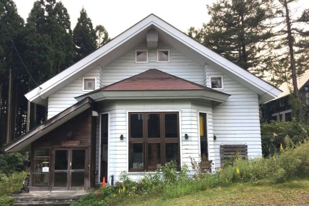 深い森を背景に立つ、白い板張り壁の木造2階建て住宅風の建物。三角屋根が特徴的で、手前には草が生い茂る斜面がある。
