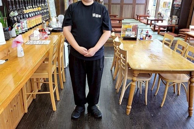 黒いTシャツの制服を着たスタッフが立つ居酒屋の店内。左側には一升瓶が並ぶカウンター席、右側には木製のテーブル席があり、奥には座敷スペースが広がっている。