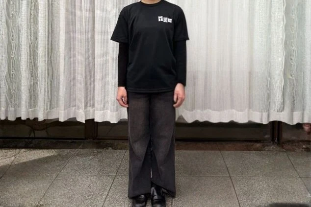 左胸に白いロゴが入った黒いTシャツと、黒いパンツを着用したスタッフの制服。背景には白いカーテンがあり、シンプルで落ち着いた印象の装い。