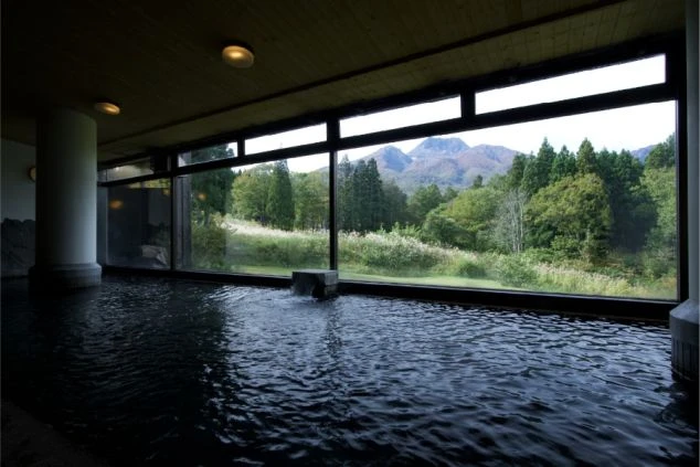 広々とした屋内の石造り大浴場。壁一面の大きな窓からは、青空の下に広がる緑豊かな森と、遠くにそびえる壮大な山々の景色を一望できる。