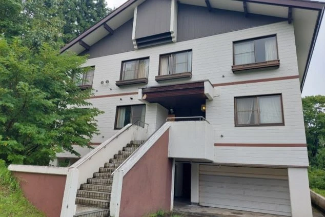 階段が特徴的な白い3階建ての建物。周辺には豊かな緑が広がり、1階部分にはガレージがある。落ち着いた雰囲気の集合住宅の外観。