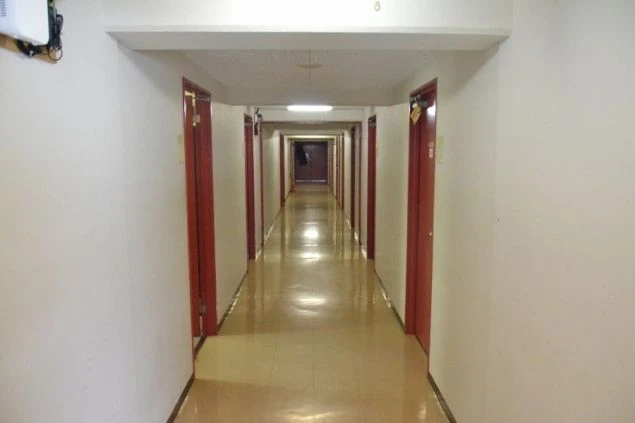 寮の長い廊下。等間隔に赤いドアの個室が並んでおり、突き当たりにもドアが見える。天井には照明があり、明るく整った印象。