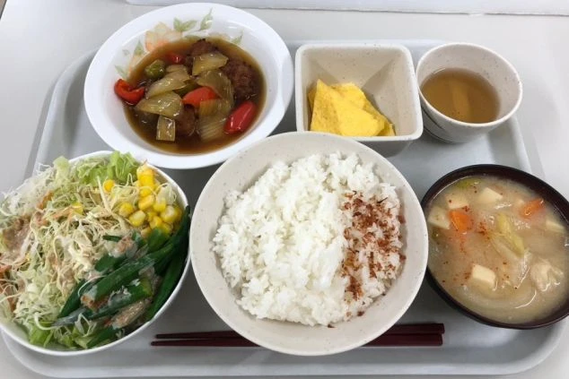 トレイに載った賄い料理。白米、味噌汁、肉と野菜の甘酢あんかけ、卵焼き、サラダ、お茶が並んでいる。