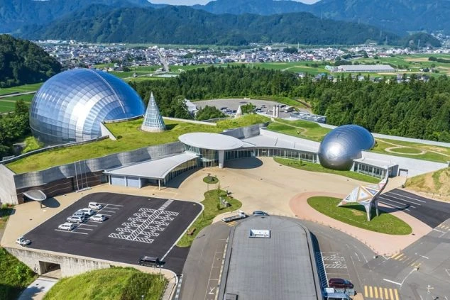 山々に囲まれたモダンな博物館の全景。銀色の巨大な球体やドーム状の建物が特徴的で、広々とした敷地内には恐竜を模したモニュメントや駐車場が整備されている。