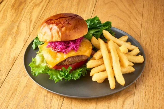 バンズに恐竜の焼印が押されたボリューム満点のハンバーガー。厚みのあるパテにチーズ、紫キャベツのマリネ、レタスが挟まれ、たっぷりのフライドポテトが添えられている。