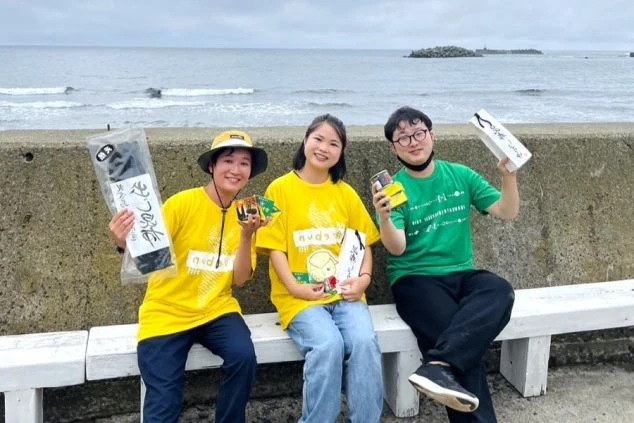 曇り空の海辺で、お土産品を手に持って笑顔で座る若者3人のグループ。黄色と緑のTシャツを着ており、和気あいあいとした旅の記念写真。