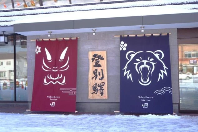 R登別駅の入り口に掲げられた鬼とクマのイラスト入り暖簾と木製の駅名看板。北海道らしい温泉地の玄関口