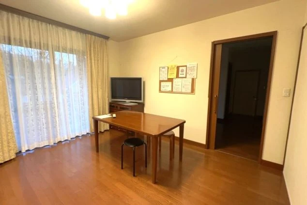 フローリングの共用リビング。中央に木製のダイニングテーブルと椅子があり、奥のテレビ台には大型テレビが設置されている。窓には白いレースのカーテンがかかっている。